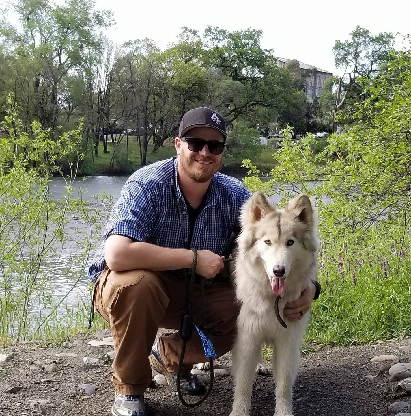 Person kneeling beside a large fluffy dog, with greenery and a river in the background.