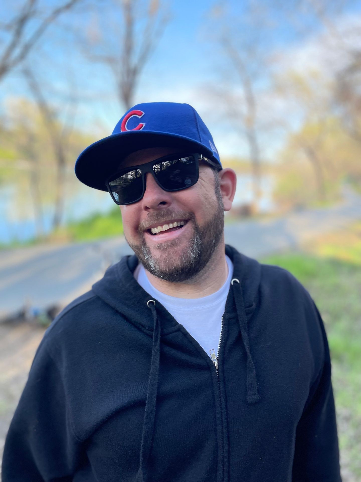 Man in sunglasses and a blue cap smiling outdoors on a sunny day.